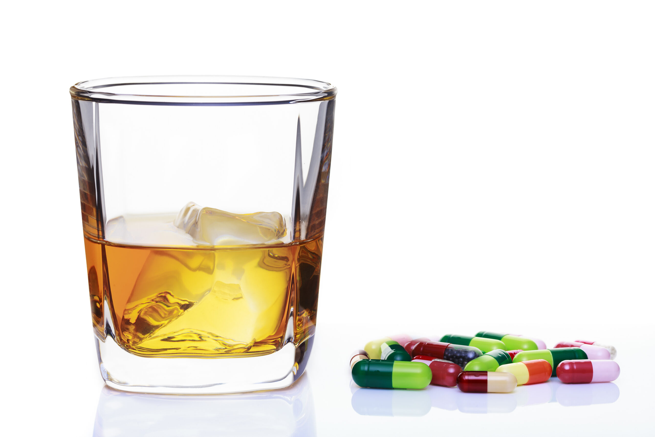 A glass of whiskey with ice beside scattered colorful capsules on a white background, illustrating the danger of mixing alcohol and medication.